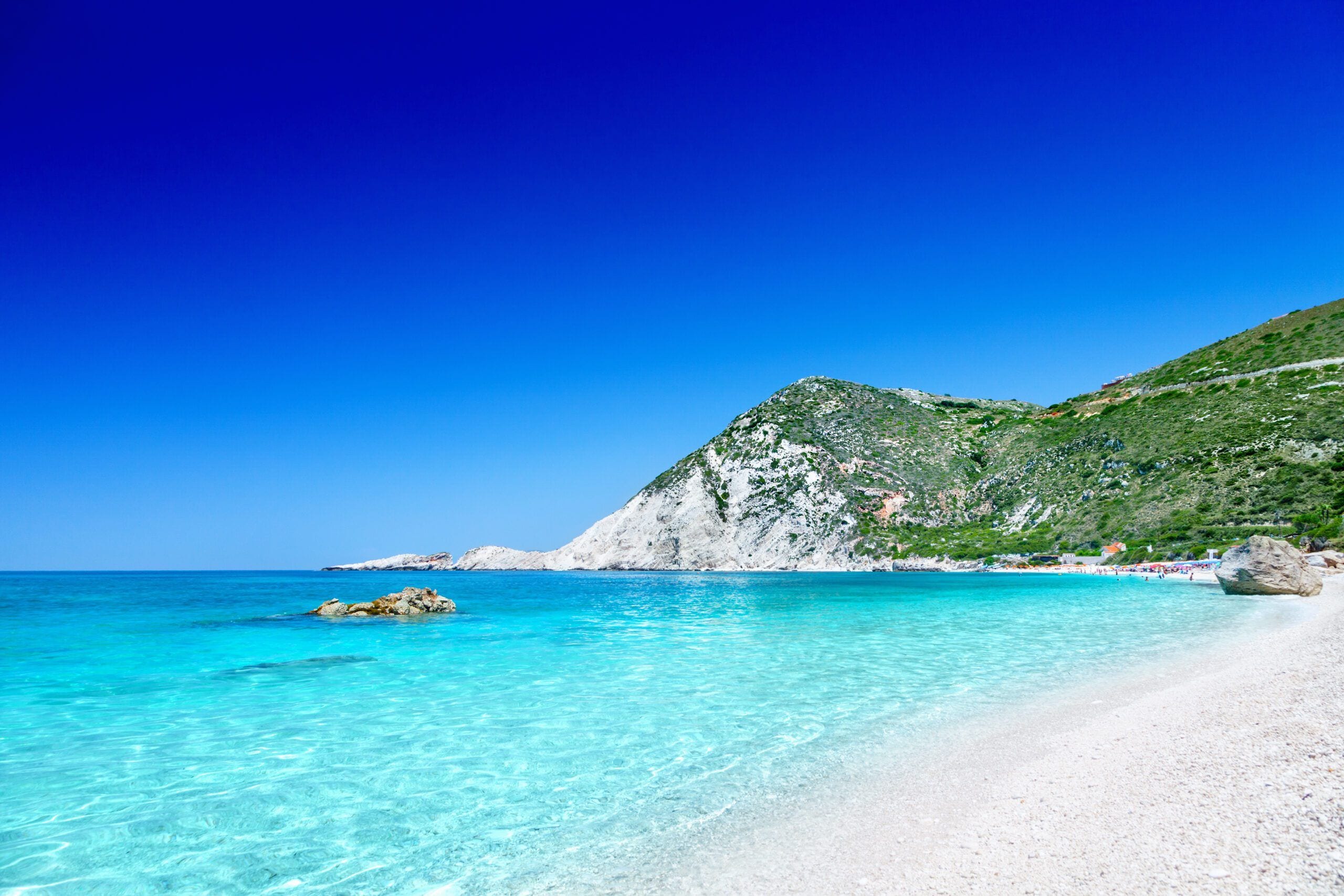 A beach with clear turquoise water, white sandy shore, and a rocky hill with greenery.