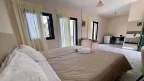 A bedroom with a large bed, a desk, and a chair, with a view of the outside through the window. - Niforos Panorama (Kefalonia)