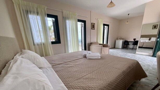 A bedroom with a large bed, a desk, and a chair, with a view of the outside through the window. - Niforos Panorama (Kefalonia)