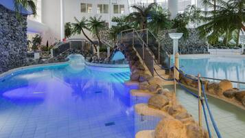An indoor swimming pool with a waterfall feature, surrounded by rocks and palm trees.