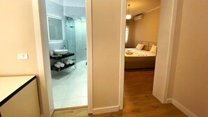 Un pasillo estrecho que conduce a un baño con cabina de ducha de vidrio y tocador con lavamanos y perchero.