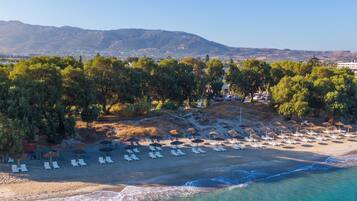 Vista aérea de una playa con filas de tumbonas y sombrillas, rodeada de árboles y montañas al fondo.