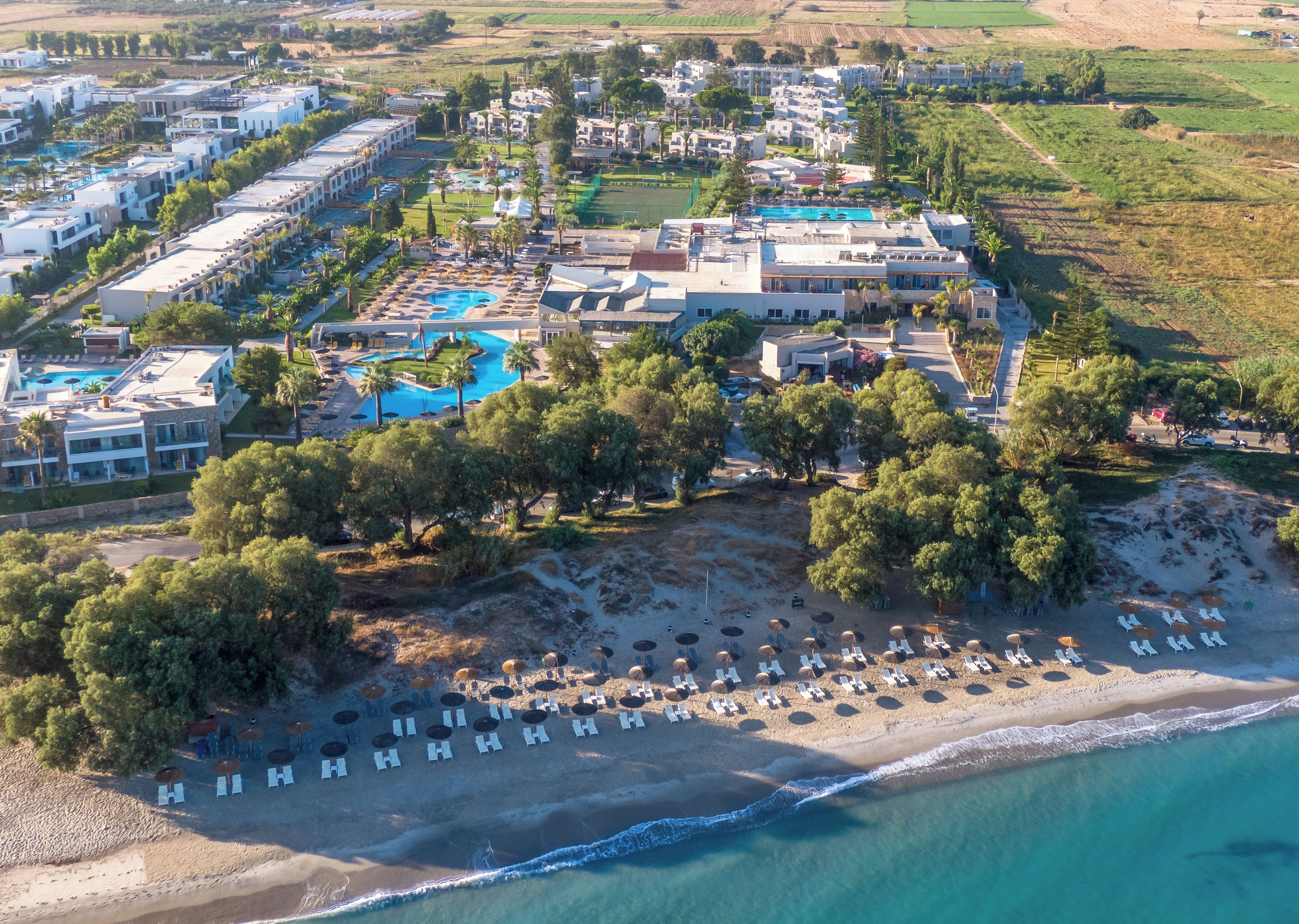 Vista aérea de un complejo turístico con una piscina grande, zona de playa con tumbonas y varios edificios.