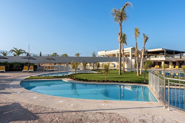 Un área de piscina con sillones de descanso, palmeras y un edificio moderno al fondo.