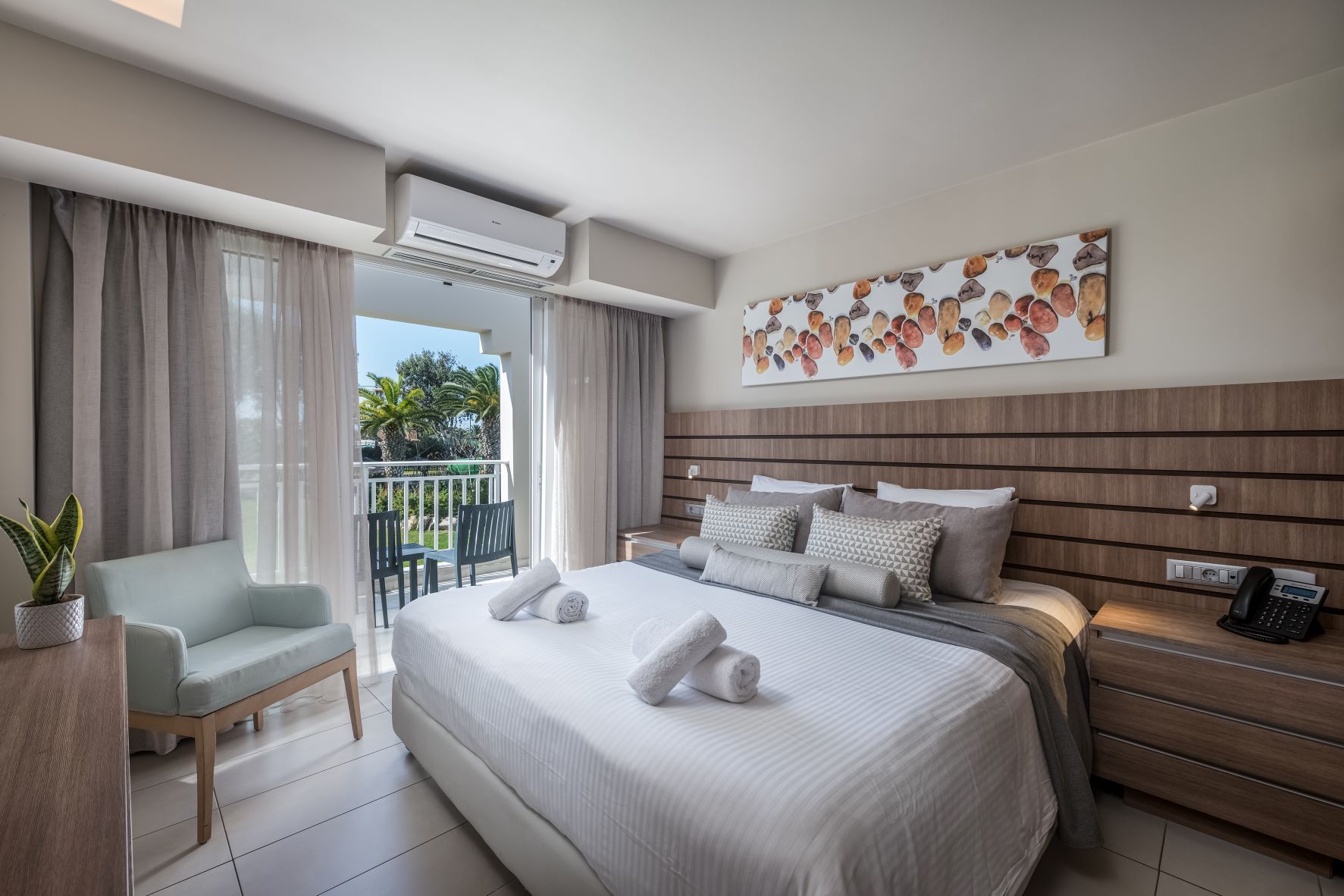 Una habitación de hotel moderna con una cama grande, un área de descanso con una silla, un balcón con cortinas y un aire acondicionado instalado en la pared.