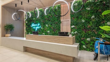 Un mostrador de recepción con detalles de madera, un letrero de neón y una pared verde decorada con plantas.