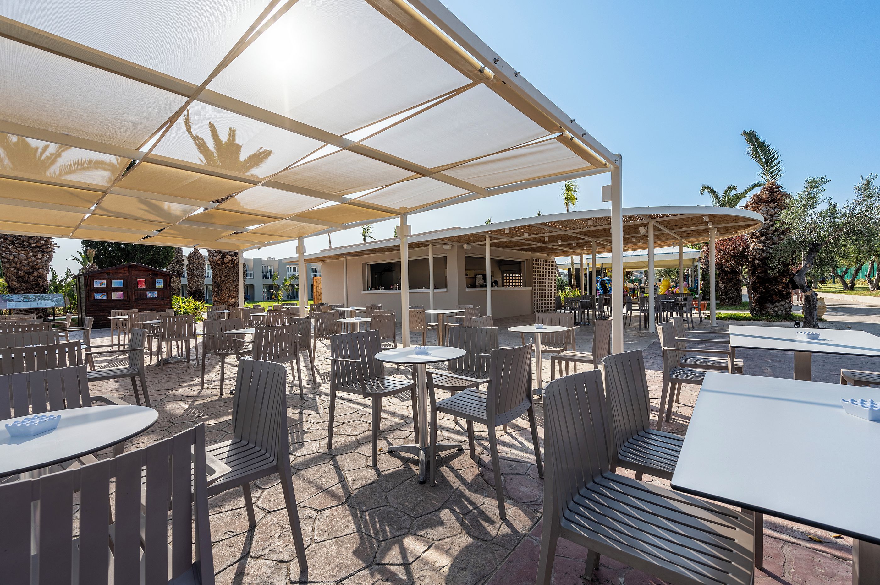 Un café al aire libre con mesas y sillas bajo una zona sombreada, un edificio al fondo y palmeras.
