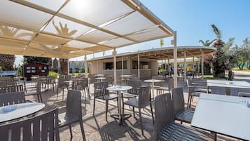 Un café al aire libre con mesas y sillas bajo una zona sombreada, un edificio al fondo y palmeras.