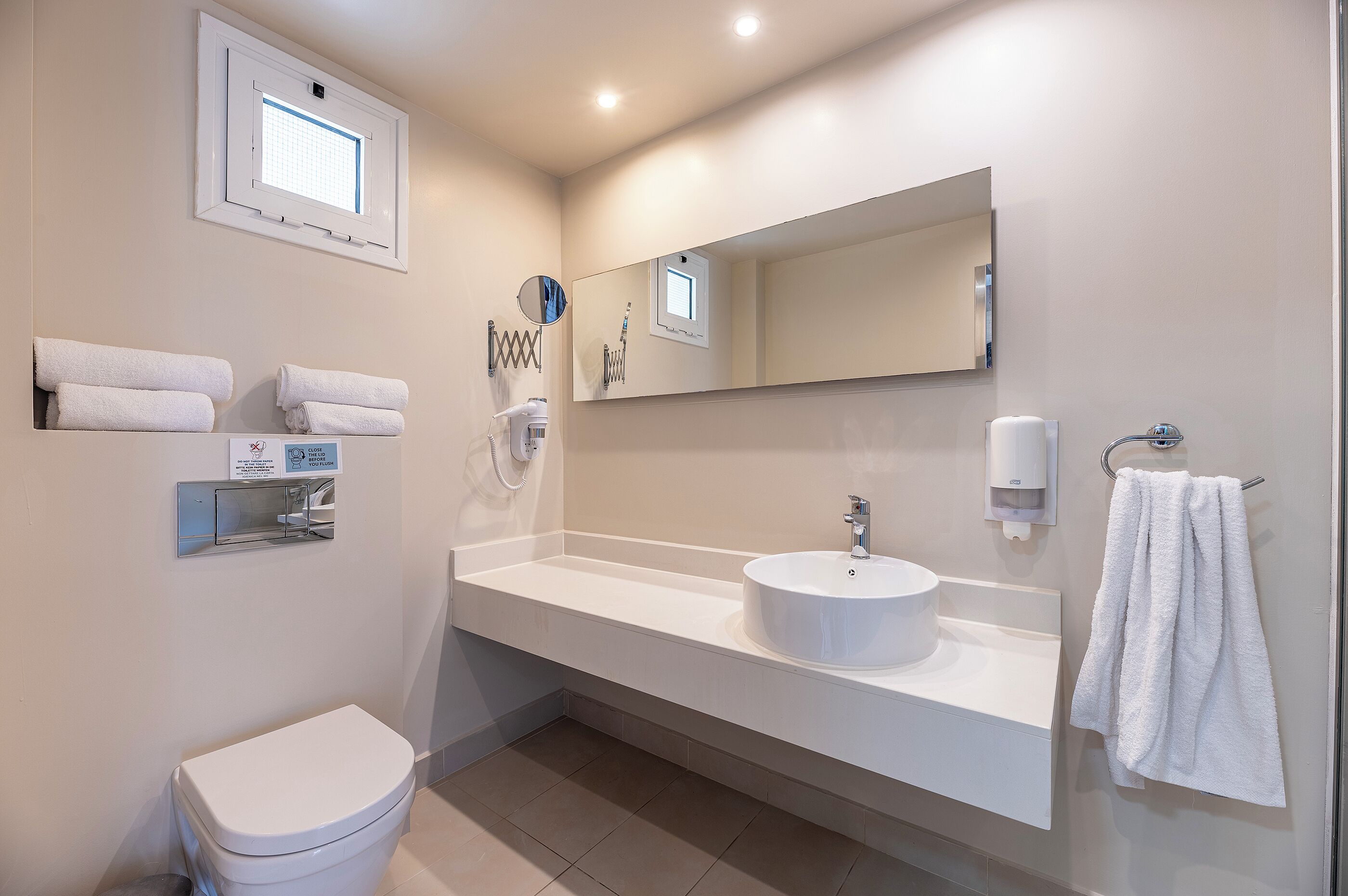 A modern bathroom with a toilet, sink, and mirror. There are towels on the wall and a window above the toilet.