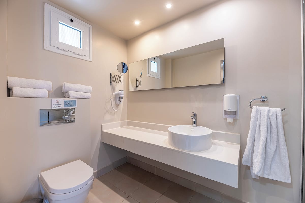 A modern bathroom with a toilet, sink, and mirror. There are towels on the wall and a window above the toilet.