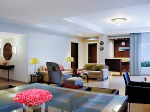 A modern hotel room with a glass dining table, two sofas, a coffee table, a lamp, and a view of the city.