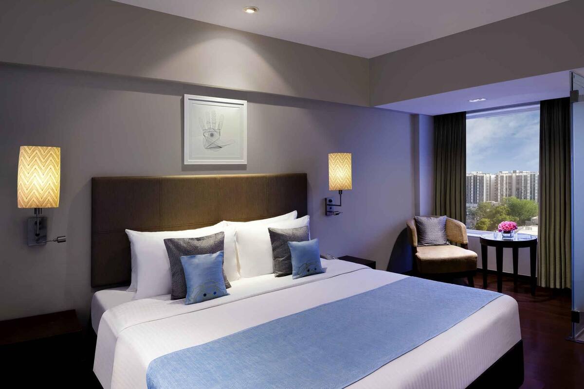 Ein Hotelzimmer mit einem großen Bett, zwei Wandlampen, einem gerahmten Kunstwerk, einem kleinen Tisch mit einer Vase und Blick auf Gebäude durch das Fenster.
