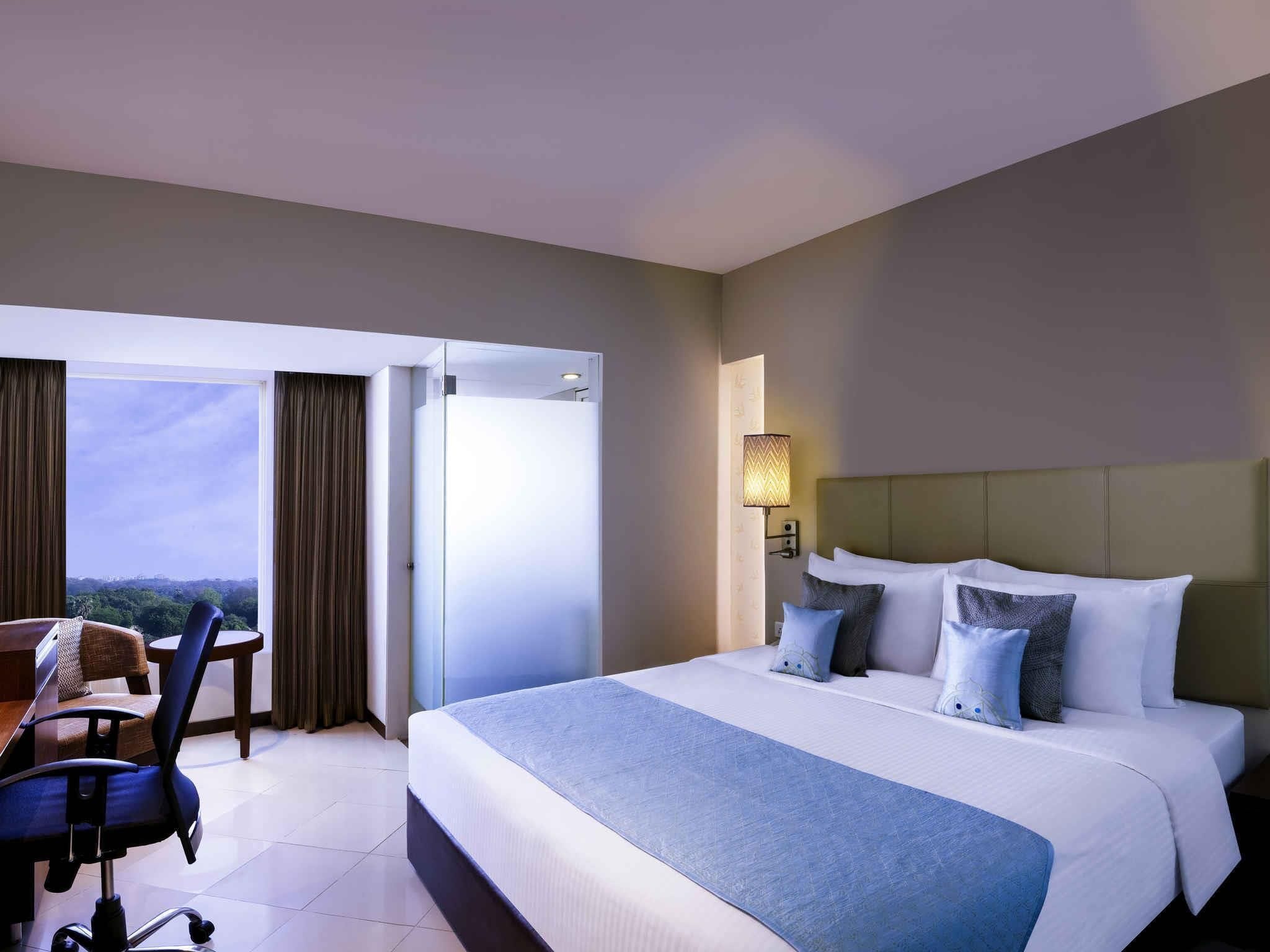Habitación de hotel con cama, escritorio y silla. Tiene una ventana con cortinas, una lámpara de pared y vistas al exterior.