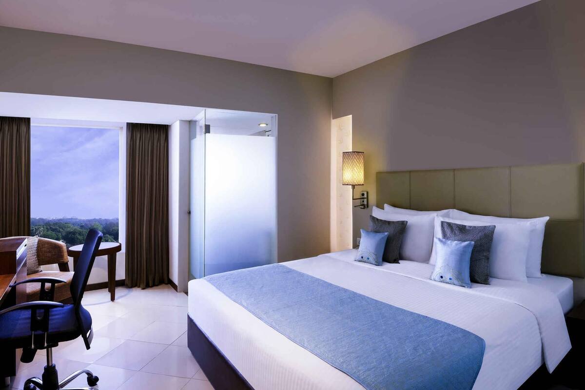 Ein Hotelzimmer mit einem Bett, einem Schreibtisch und einem Stuhl. Es gibt ein Fenster mit Vorhängen, eine Wandlampe und einen Blick ins Freie.