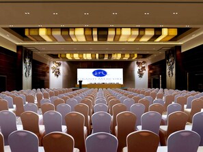 Uma sala de conferências com fileiras de cadeiras, um palco com tela de apresentação e um design de teto moderno.