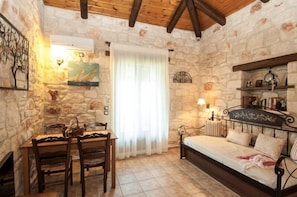 A stone-walled room with a wooden ceiling, a dining table with chairs, a bed with a headboard, and a window with a white curtain.