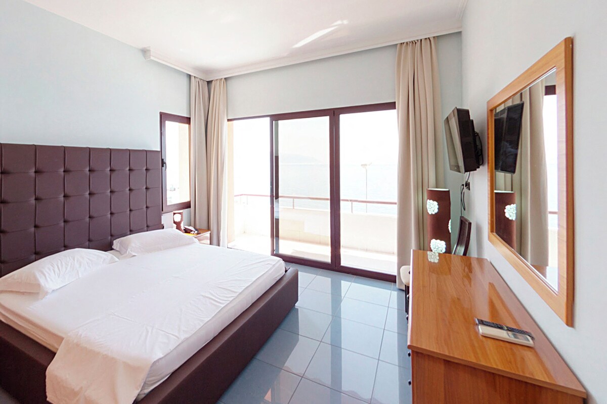 Habitación de hotel con una cama grande, un escritorio de madera, un televisor y un balcón con cortinas.