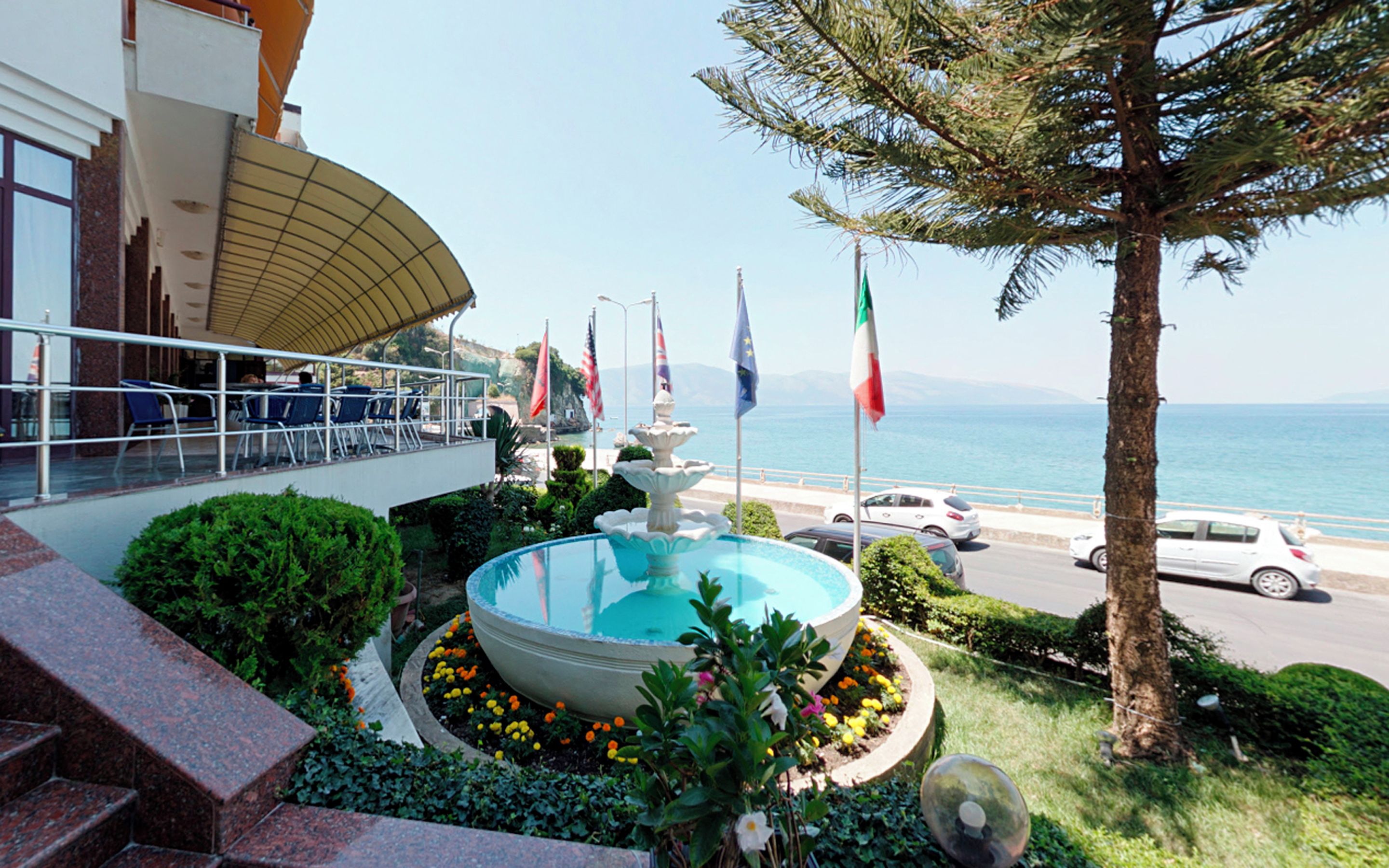 A pool with a fountain, surrounded by flowers and flags, overlooking a coastal road with cars.