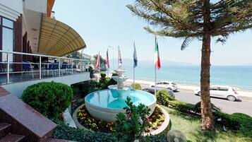 A pool with a fountain, surrounded by flowers and flags, overlooking a coastal road with cars.