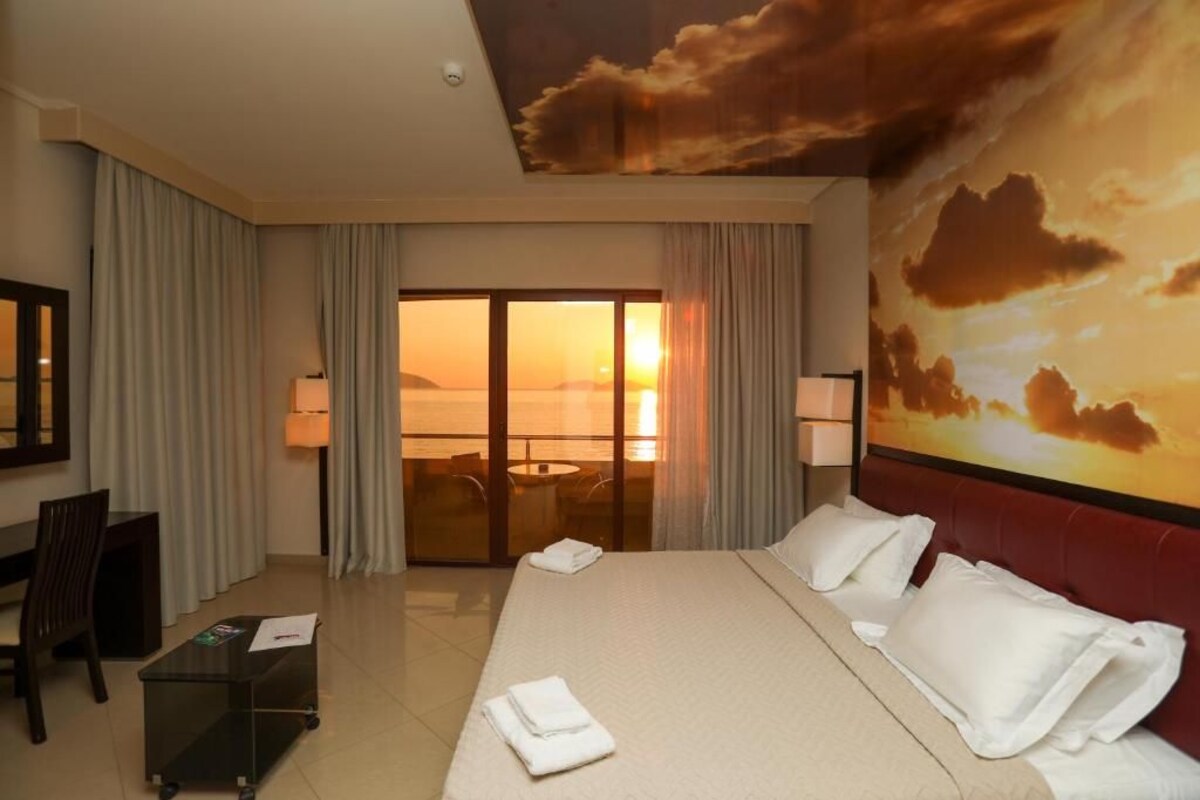 Habitación de hotel con cama, escritorio, silla, vista al atardecer por la ventana y un cuadro de nubes en el techo.