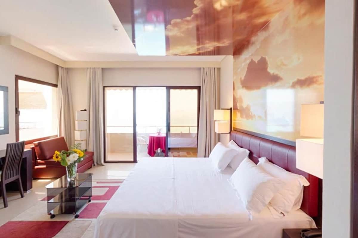 A hotel room with a large bed, a sofa, a desk, and a balcony with a view.
