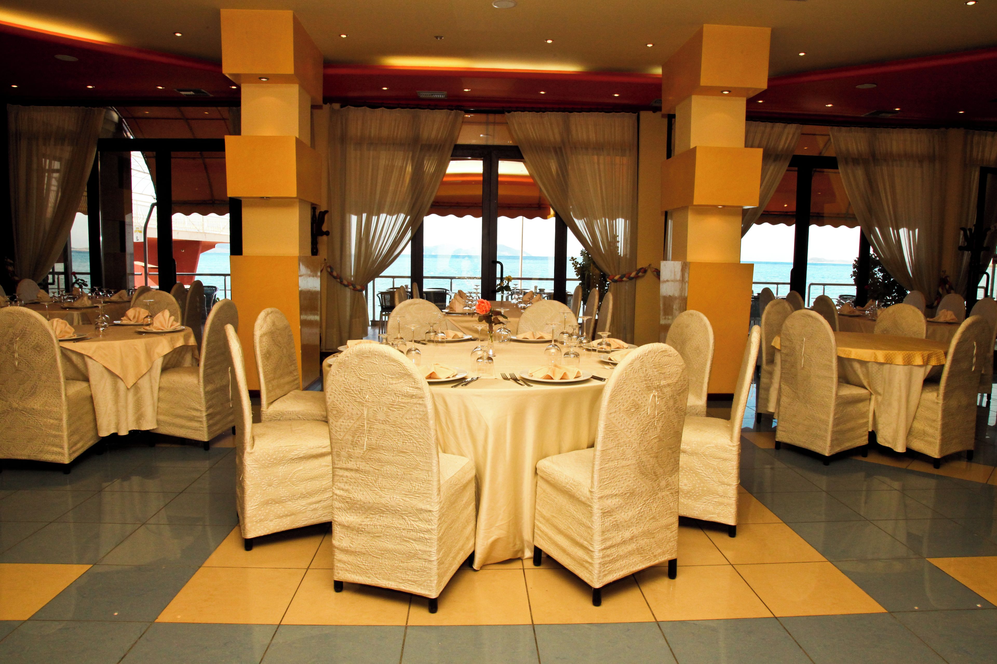 Un espace repas avec des tables et des chaises rondes, de grandes fenêtres avec des rideaux et une vue sur la mer.