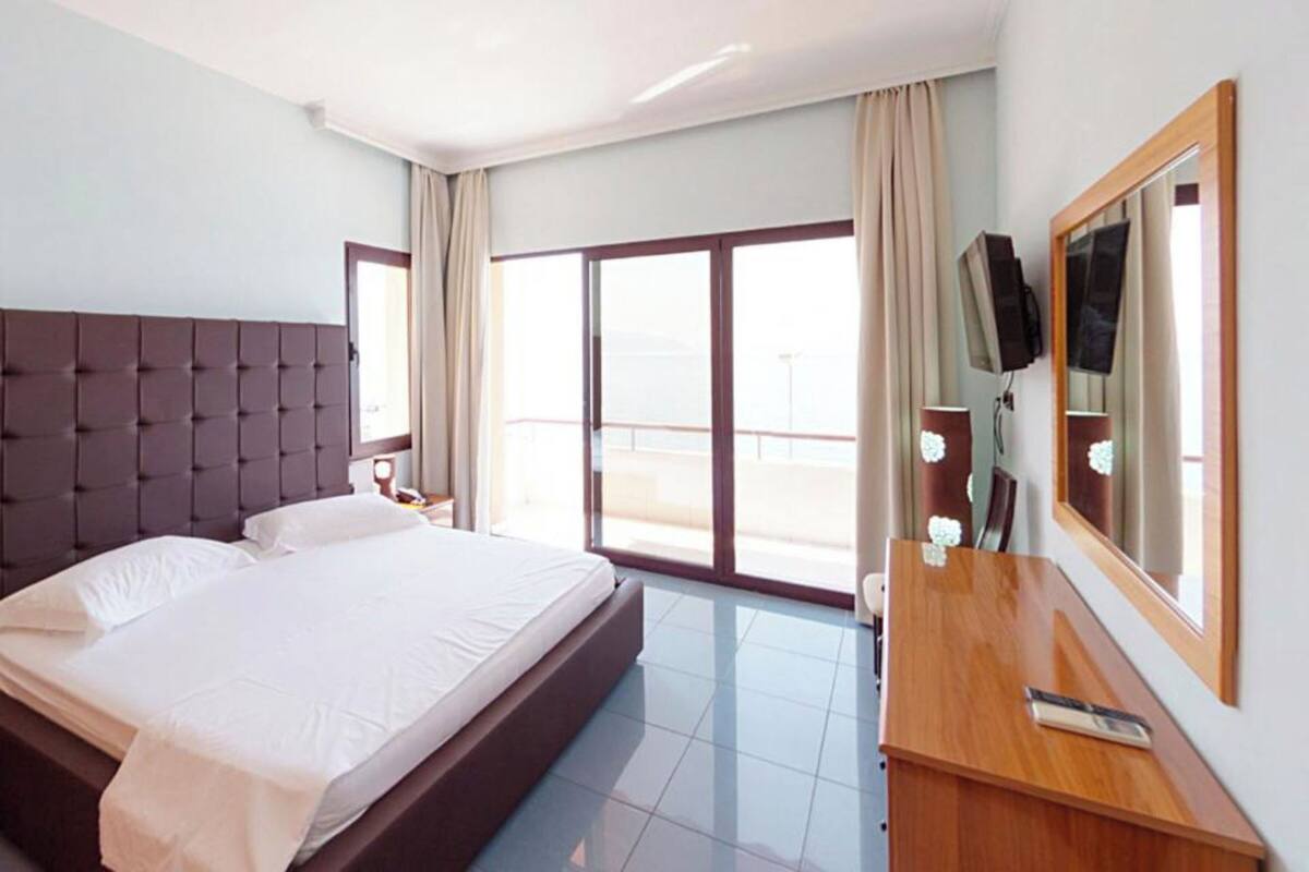Habitación de hotel con una cama grande, un escritorio de madera, un televisor y un balcón con cortinas.