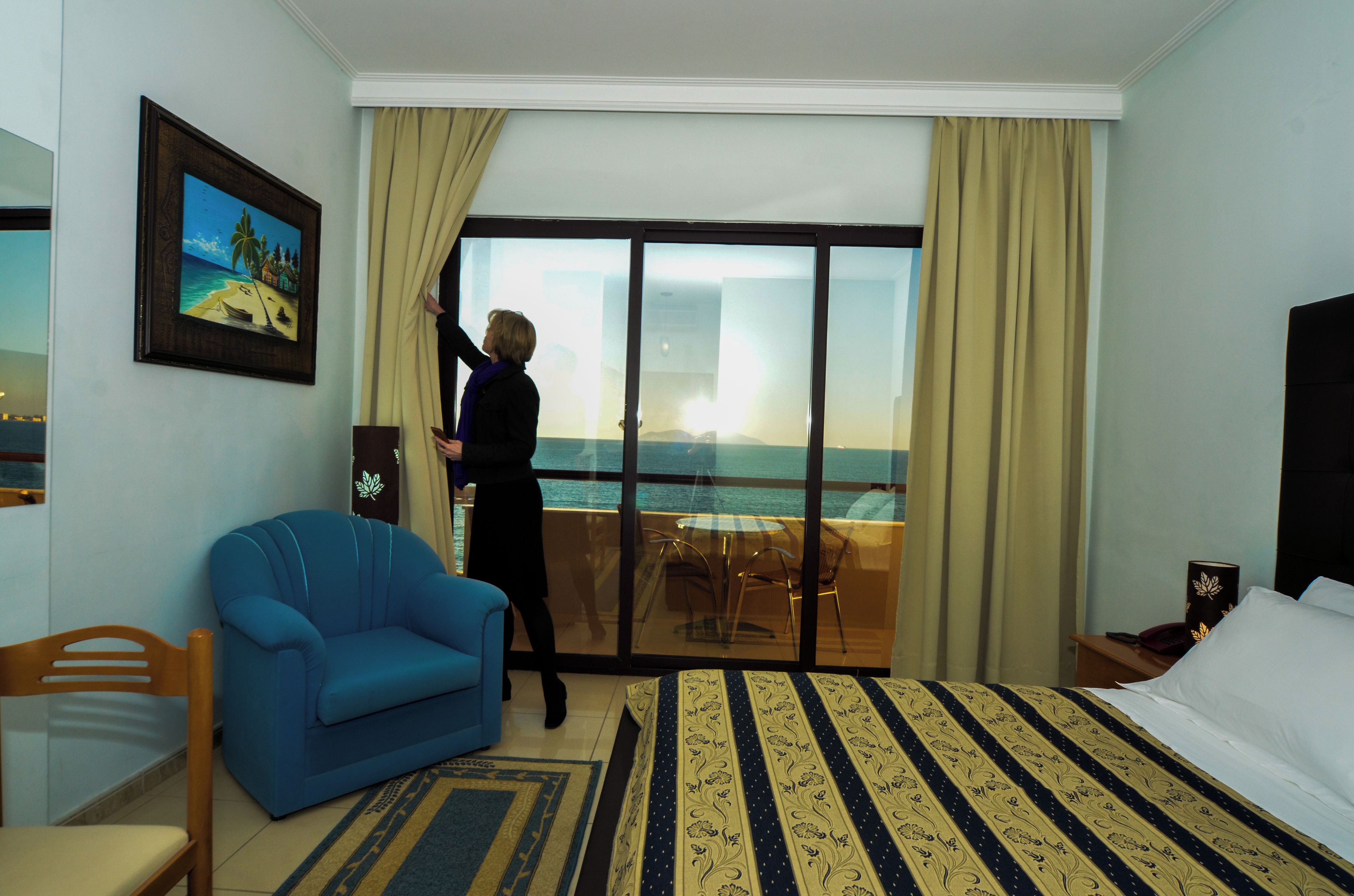 A hotel room with a large bed, a blue armchair, a dining area with a table and chairs, and a view of the sea through a glass door.