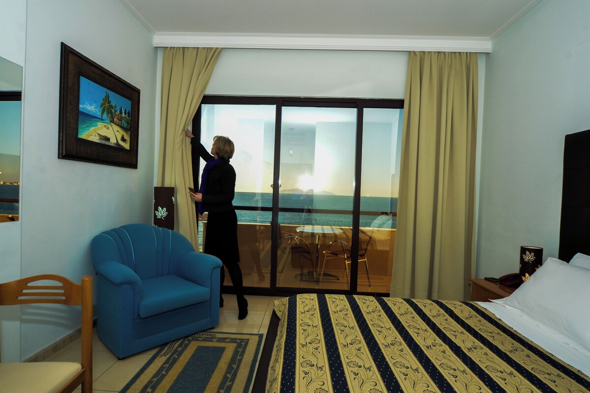 Habitación de hotel con una cama grande, un sillón azul, un área de comedor con mesa y sillas, y vista al mar a través de un ventanal.