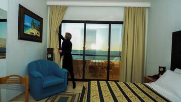 A hotel room with a large bed, a blue armchair, a dining area with a table and chairs, and a view of the sea through a glass door.