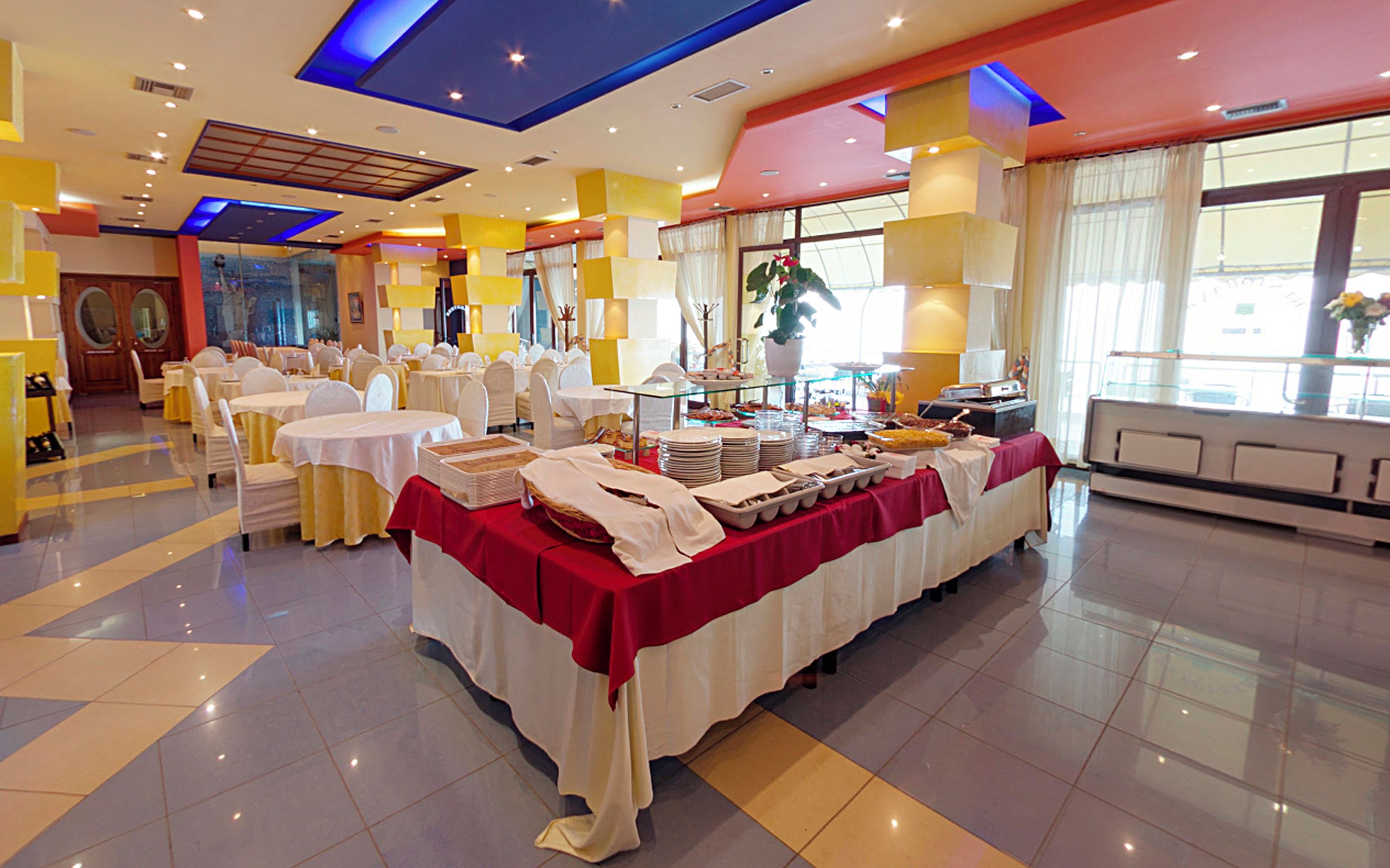 A banquet hall with round tables, a buffet setup, and a large window providing natural light.