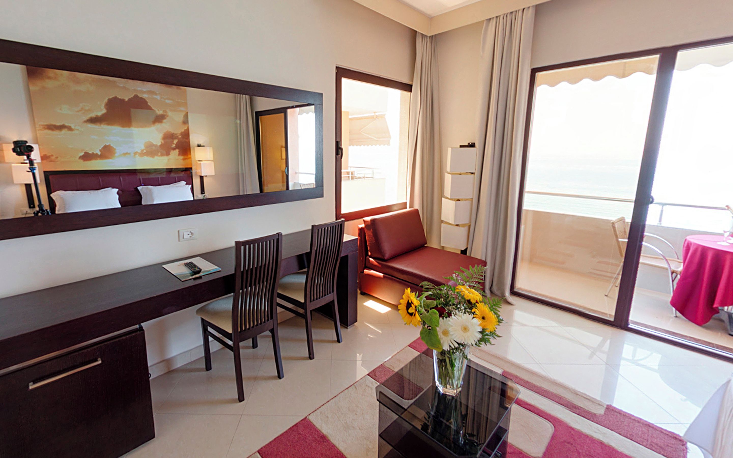A hotel room with a desk, chairs, a sofa, a large window, and a balcony with a table.