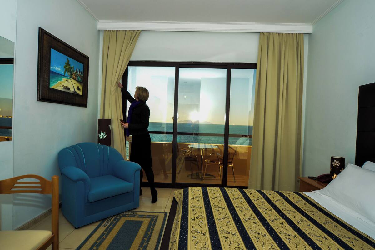 Habitación de hotel con una cama grande, un sillón azul, una mesita pequeña y vista al océano a través de un ventanal corredero.