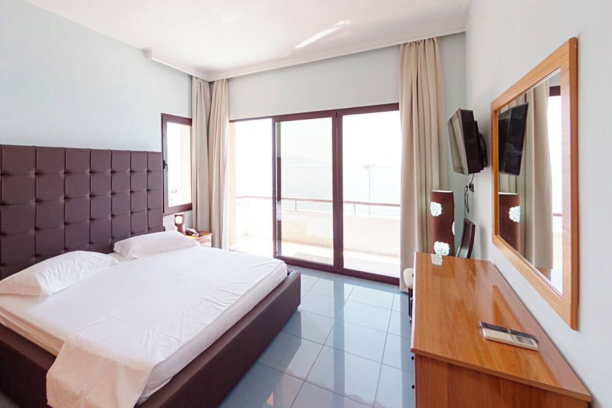 Habitación de hotel con una cama grande, un escritorio de madera, un televisor y un balcón con cortinas.
