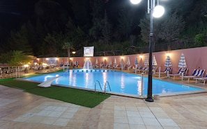 A swimming pool with a fountain, surrounded by lounge chairs and umbrellas, with a signboard in the background.
