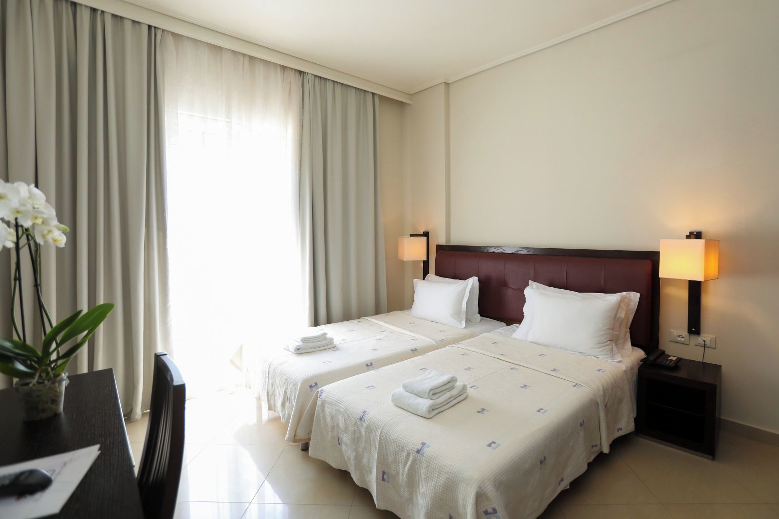 Une chambre d’hôtel avec deux lits, un bureau, une chaise, un vase de fleurs et une lampe sur la table de chevet.