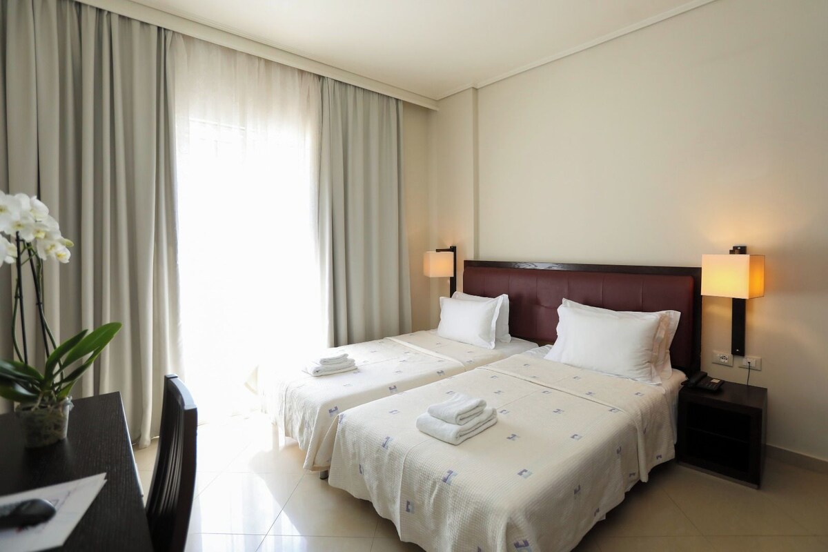 Habitación de hotel con dos camas, un escritorio, una silla, un jarrón con flores y una lámpara en la mesita de noche.