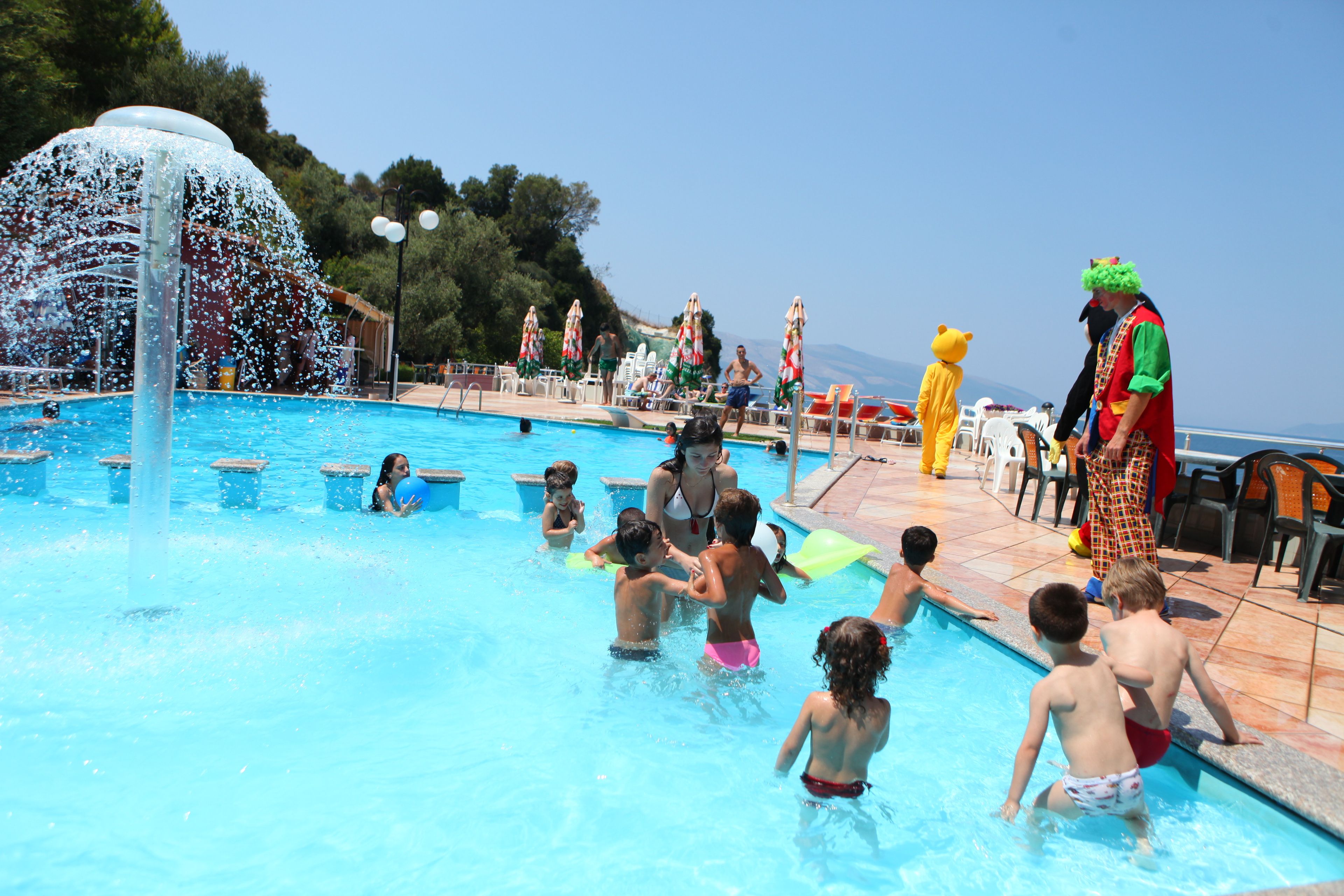A swimming pool with children and adults, some in costumes, and a water fountain feature.