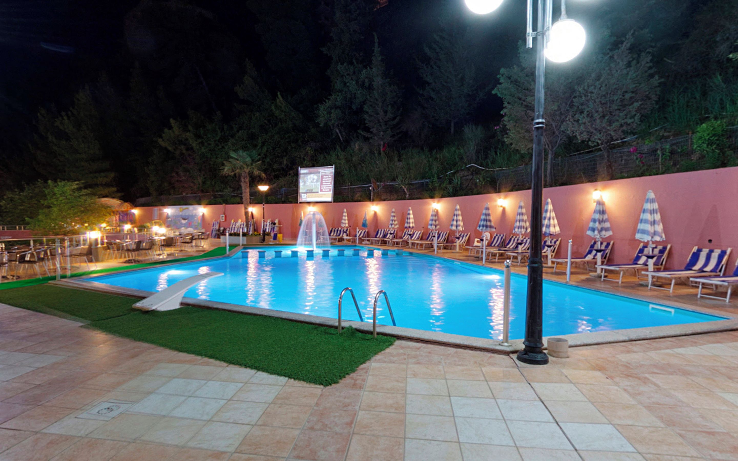 A swimming pool with a fountain, surrounded by lounge chairs and umbrellas, with a signboard in the background.
