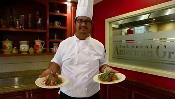 A chef in a white uniform holding two plates of food in a kitchen with red walls and wooden cabinets. - Hilton Garden Inn Fayetteville/Fort Bragg (FAYETTEVILLE)