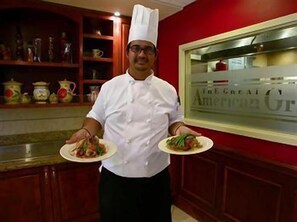 A chef in a white uniform holding two plates of food in a kitchen with red walls and wooden cabinets. - Hilton Garden Inn Fayetteville/Fort Bragg (FAYETTEVILLE)