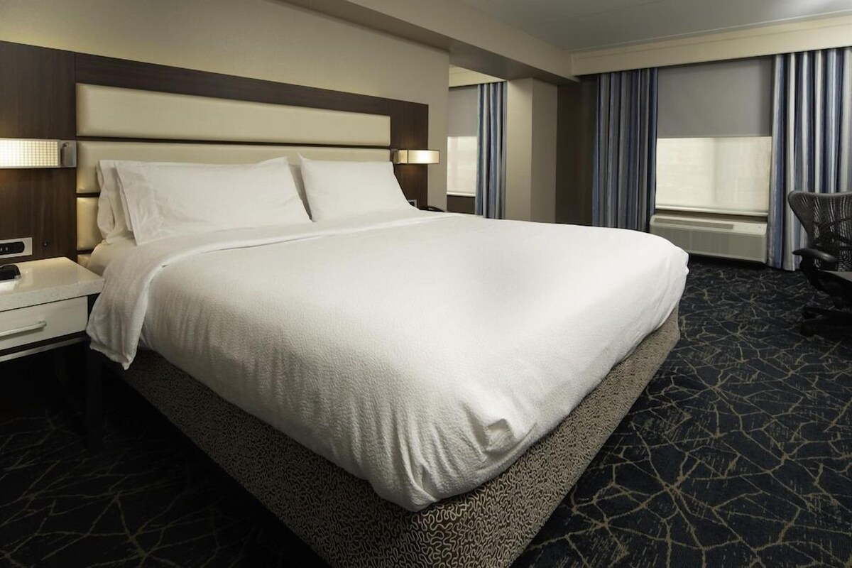 A hotel room with a large bed, a nightstand, a chair, and a window with curtains.