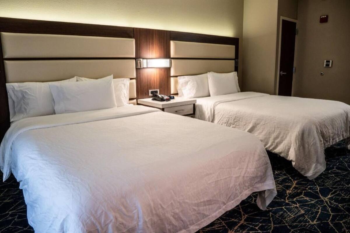 A hotel room with two beds, a nightstand with a phone, and a wall-mounted light.