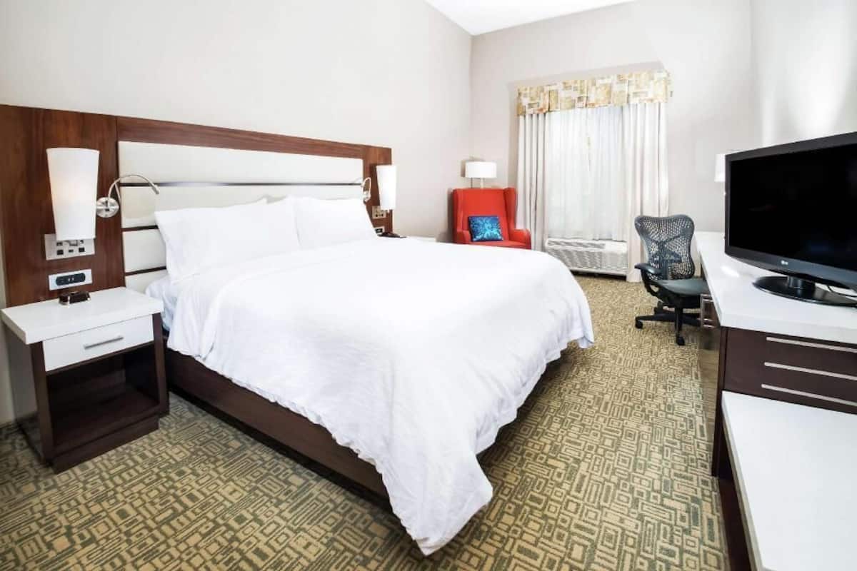 A hotel room with a bed, a desk, a television, a chair, and a nightstand with a lamp.