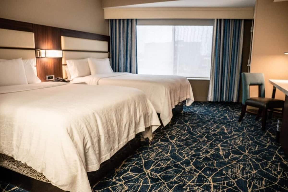 A hotel room with two beds, a desk, a chair, and a large window with curtains.