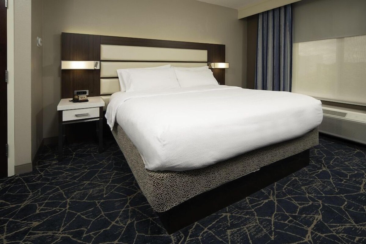 A hotel room with a bed, a nightstand, a lamp, and a window with curtains.