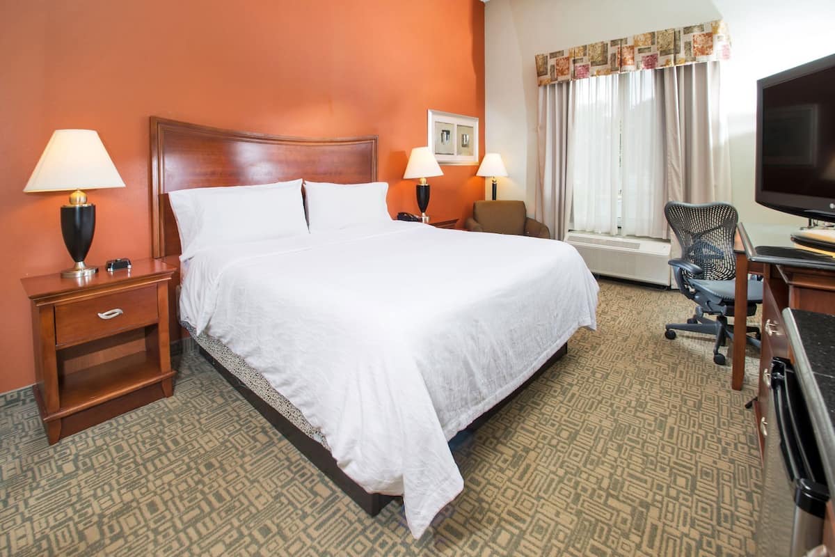 A hotel room with a large bed, two bedside tables with lamps, a desk with a chair, a television, and a window with curtains.
