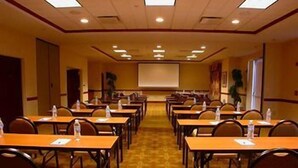 A conference room with rows of tables and chairs, a projector screen, and a carpeted floor. - Hilton Garden Inn Fayetteville/Fort Bragg (FAYETTEVILLE)