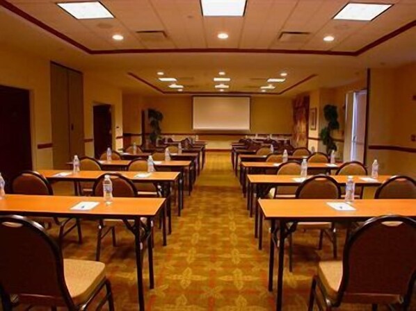 Uma sala de conferência com fileiras de mesas e cadeiras, uma tela de projetor e piso coberto de carpete.