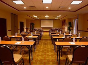 A conference room with rows of tables and chairs, a projector screen, and a carpeted floor. - Hilton Garden Inn Fayetteville/Fort Bragg (FAYETTEVILLE)